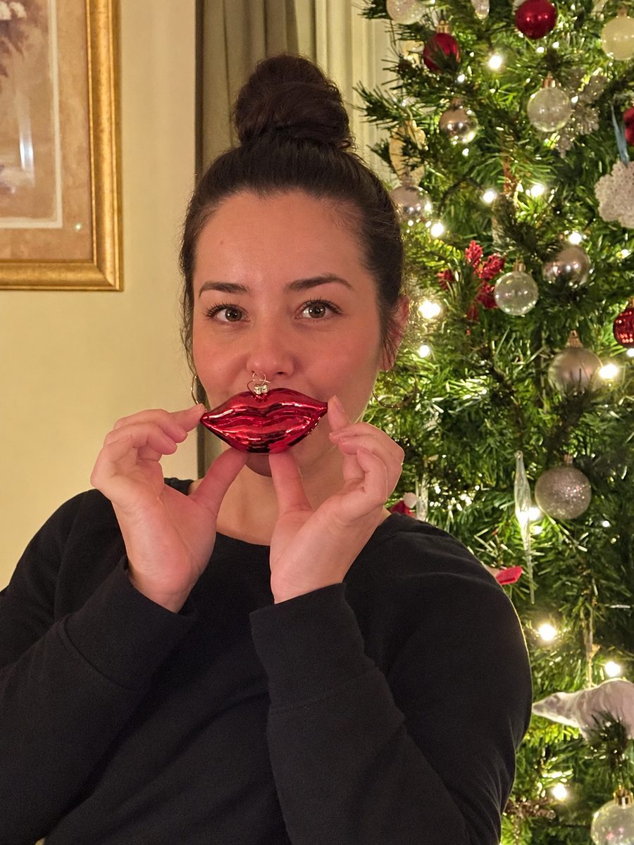 Val, Christmas decorator at Deco Designs, holding a red lips bauble in front of a lit Christmas tree
